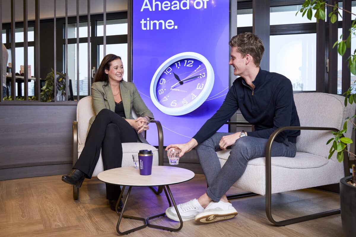 Twee professionals in gesprek bij Déhora over tijdintelligentie en strategische planning bij een wanddecoratie van een paarse klok.